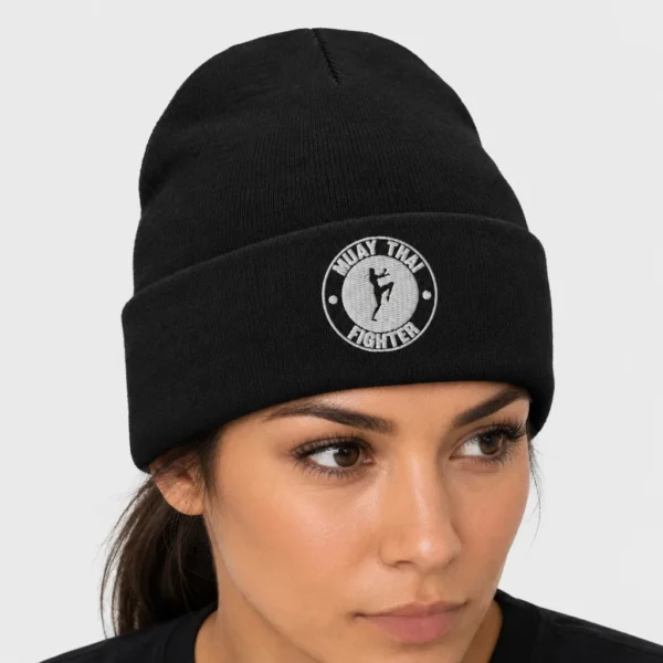 Bonnet en maille avec, brodé sur le devant, un badge circulaire montrant la silhouette d'une combattante de muay thaï genou levé entourée du texte « MUAY THAI FIGHTER ». – photo studio femme