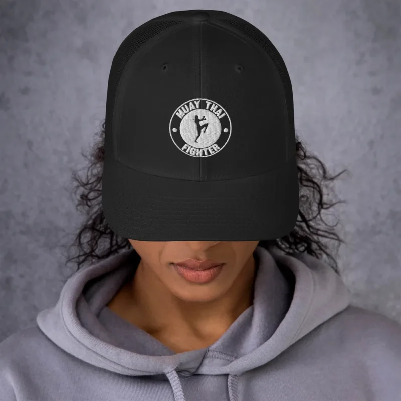 Casquette trucker rétro brodée sur le devant d'un badge circulaire avec la silhouette d'une combattante de muay-thaï genou levé et le texte « MUAY THAI FIGHTER ». (Noir, porté femme)