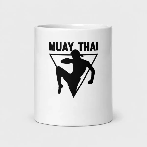 Mug blanc brillant orné de la silhouette noire d'un combattant de muay-thaï portant un mongkol, qui donne un coup de genou sauté devant un triangle inversé, sous le titre « MUAY THAI ». – photo studio